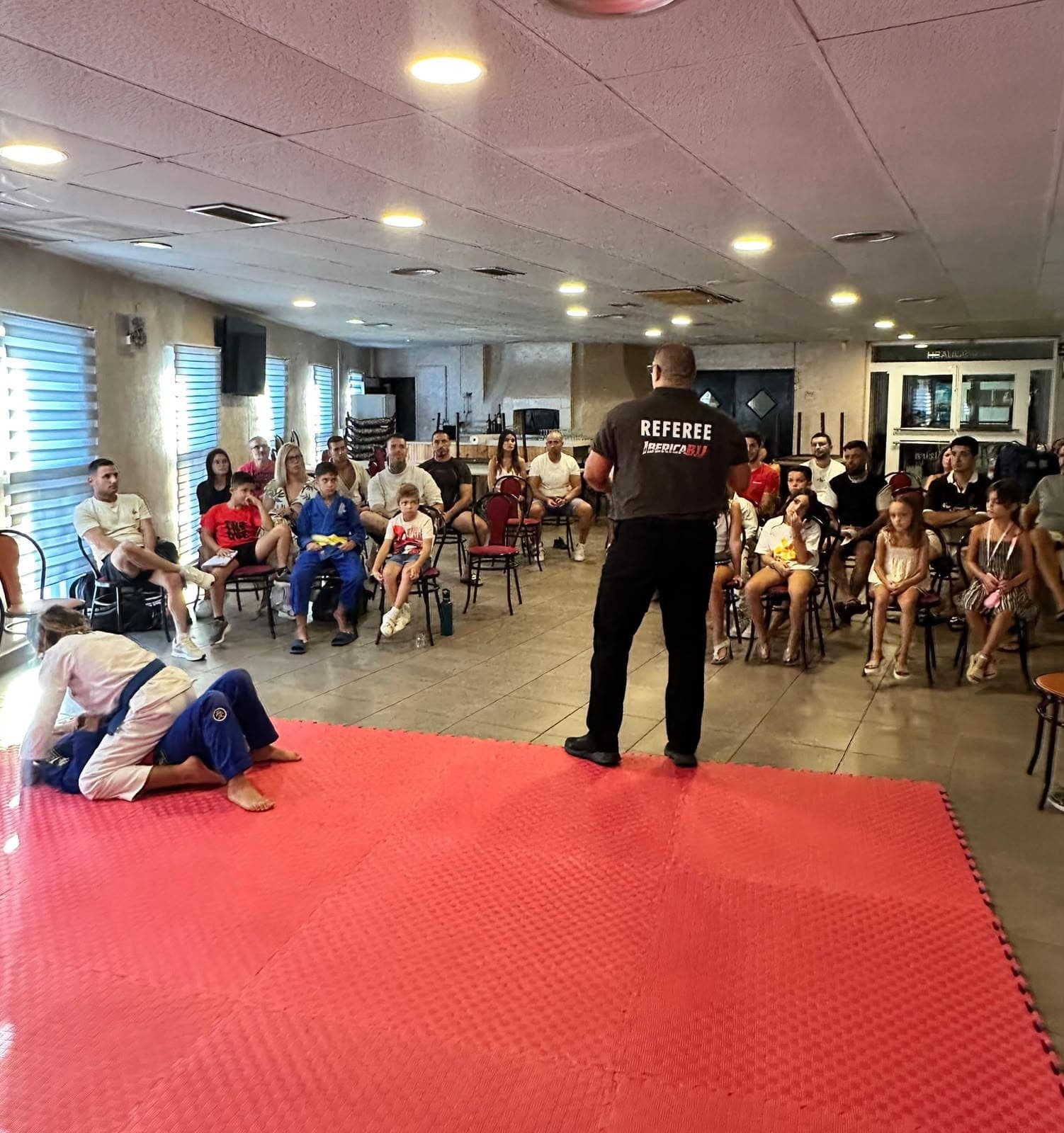 El Mestre Javier Sales, con camiseta de árbitro de Ibérica BJJ, dirige una formación ante un grupo sentado mientras dos luchadores hacen la demostración