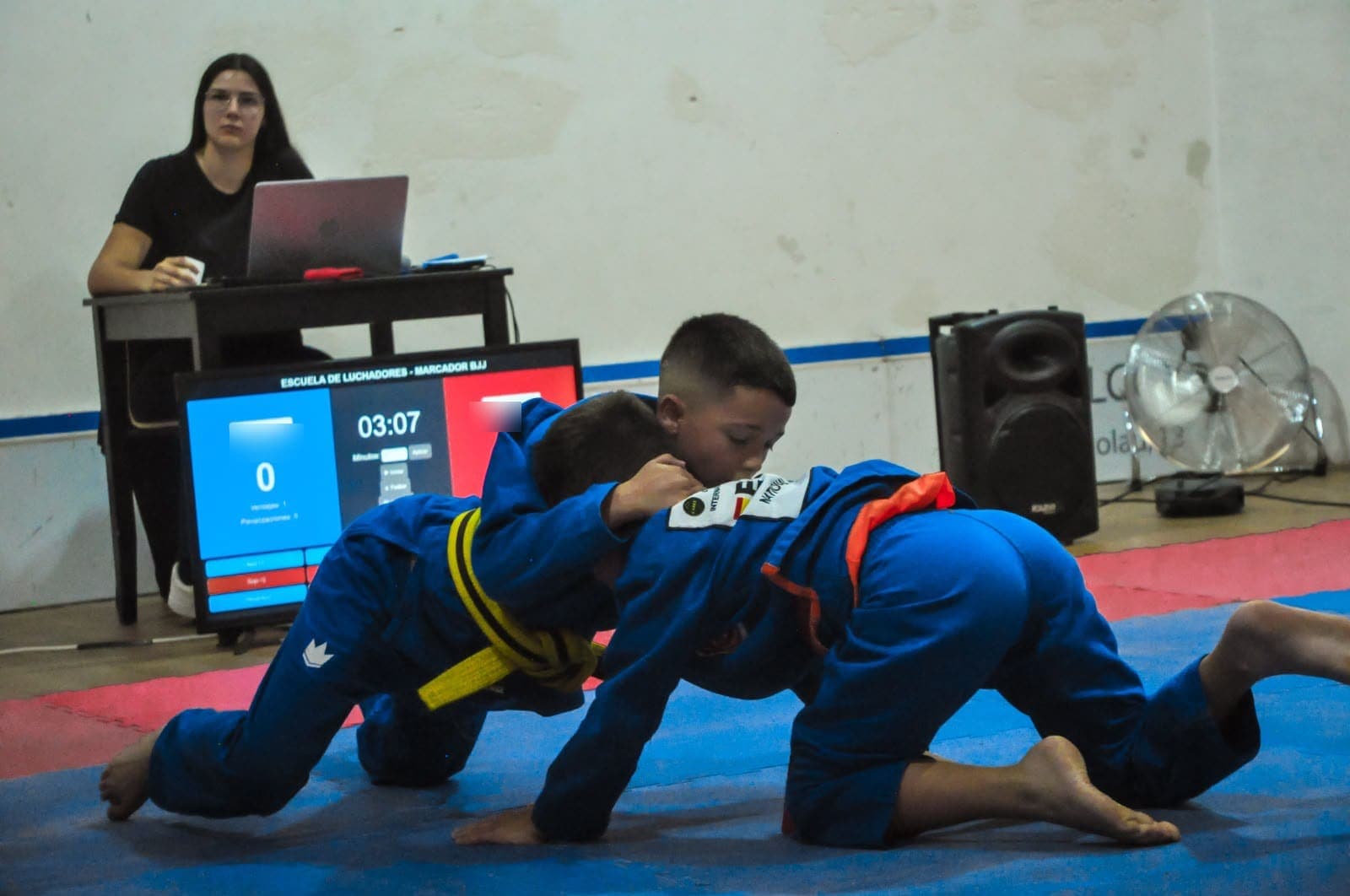 Dos niños con kimono luchan en el tatami durante una competición de la escuela, con el marcador de BJJ en pantalla